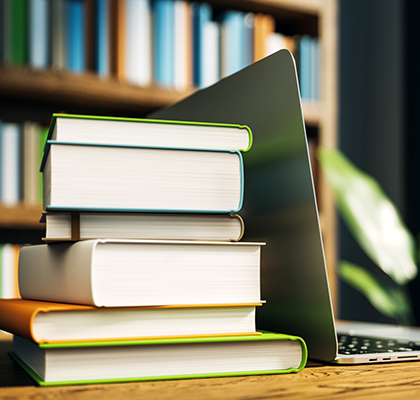 Stack of books next to an open laptop on a wooden desk in a library. Blurred bookshelf background. Concept of education and online study. 3D Rendering