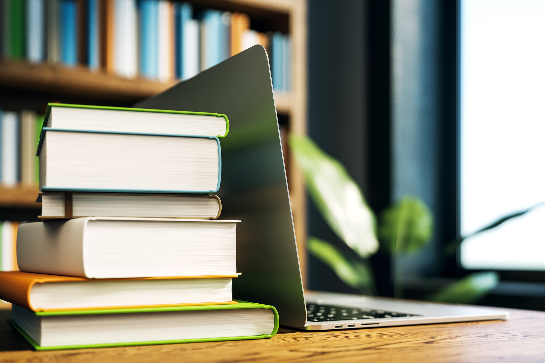 Stack of books next to an open laptop on a wooden desk in a